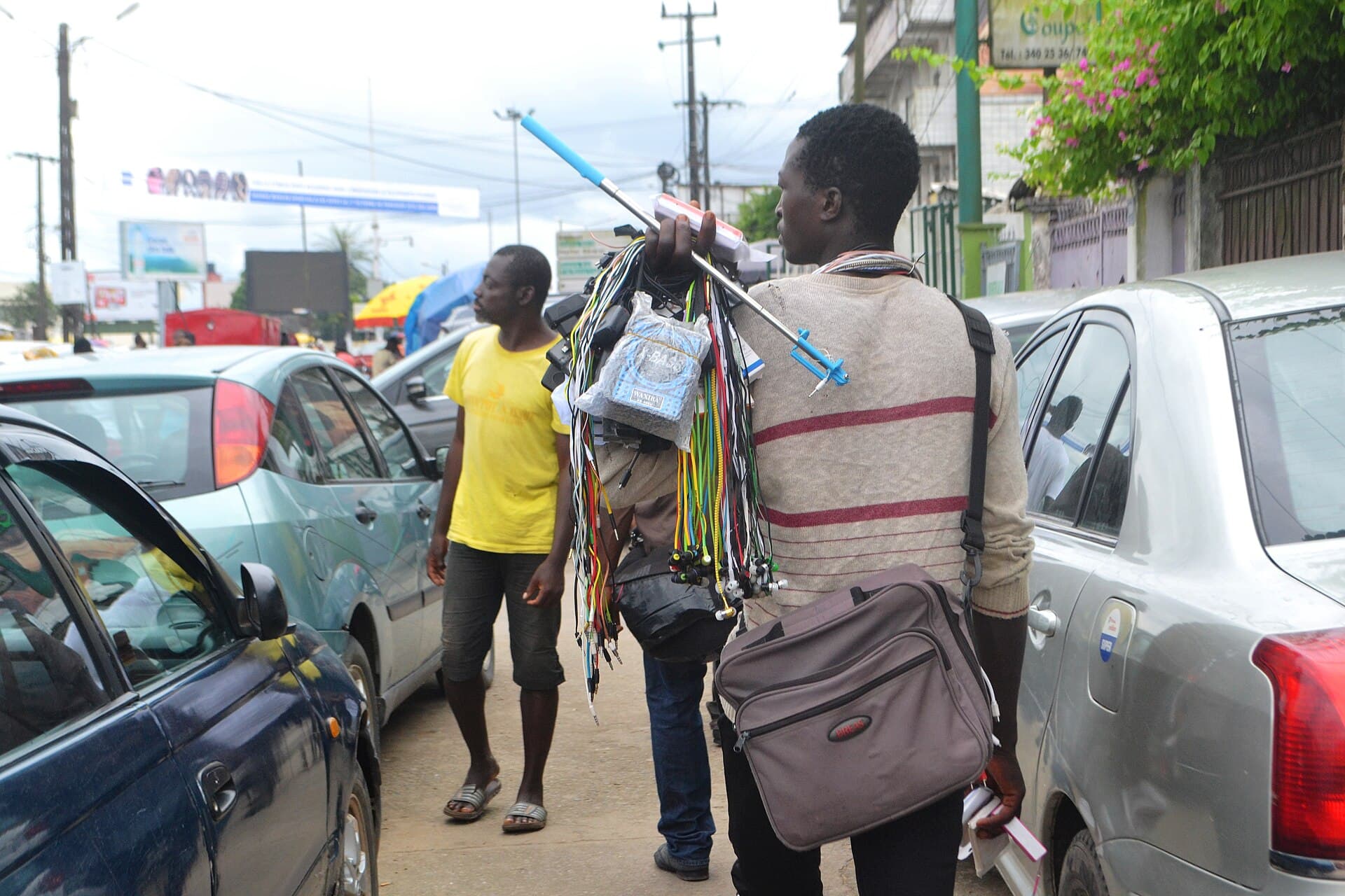 Vendeur ambulant portant en bandoulière des câbles et chargeurs de téléphone dans une rue de Douala